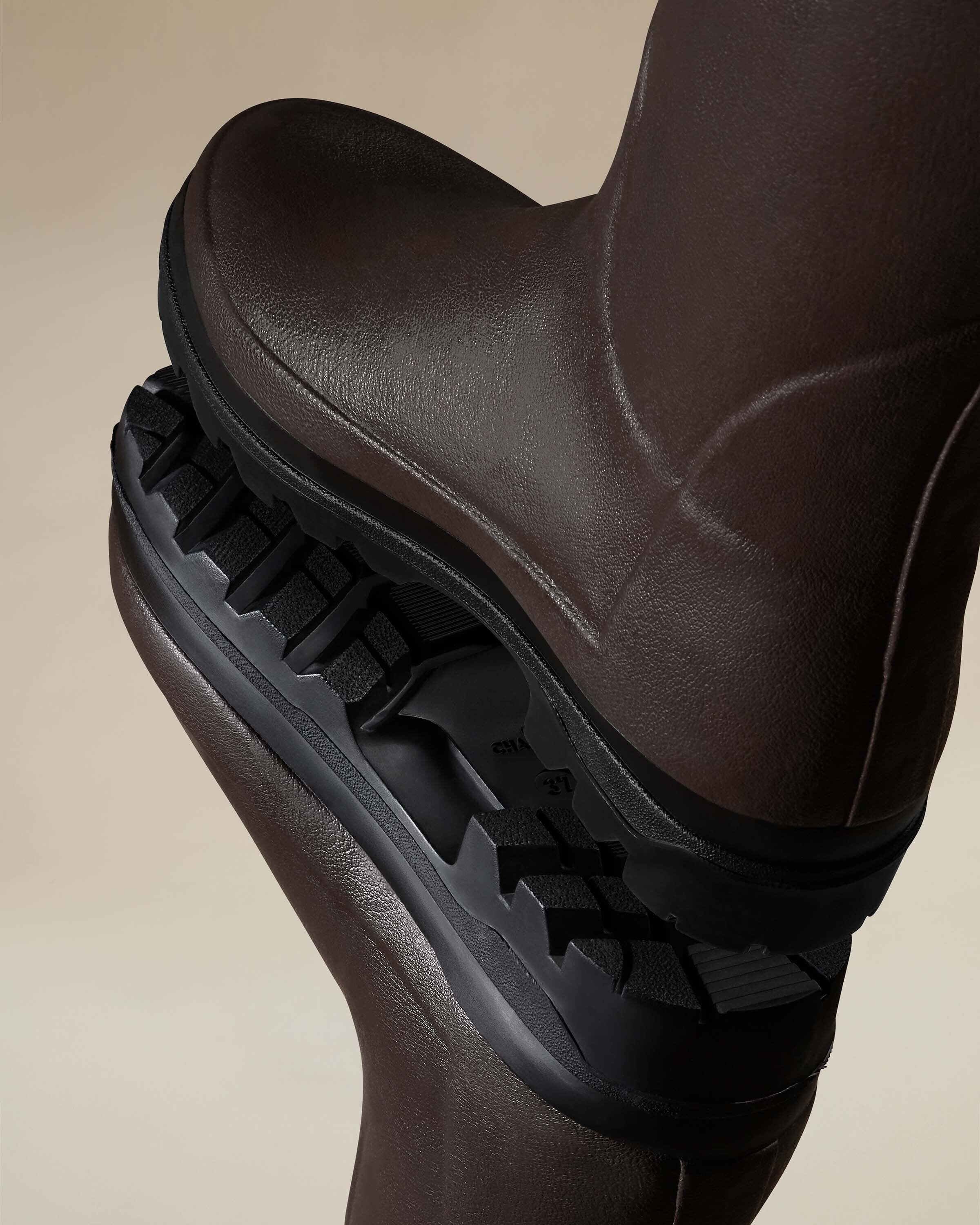 Dramatic macro image of the outsole and forefoot of the Women's Vierzonord Neoprene Lined Wellington Boot in Dark Brown, captured mid-step as one boot rests on top of the other. The aggressive lug pattern of the black sole is prominently displayed, emphasizing its suitability for uneven, wet, and slippery terrain. The image also shows the flexibility of the natural rubber as the boots bend slightly under the weight.