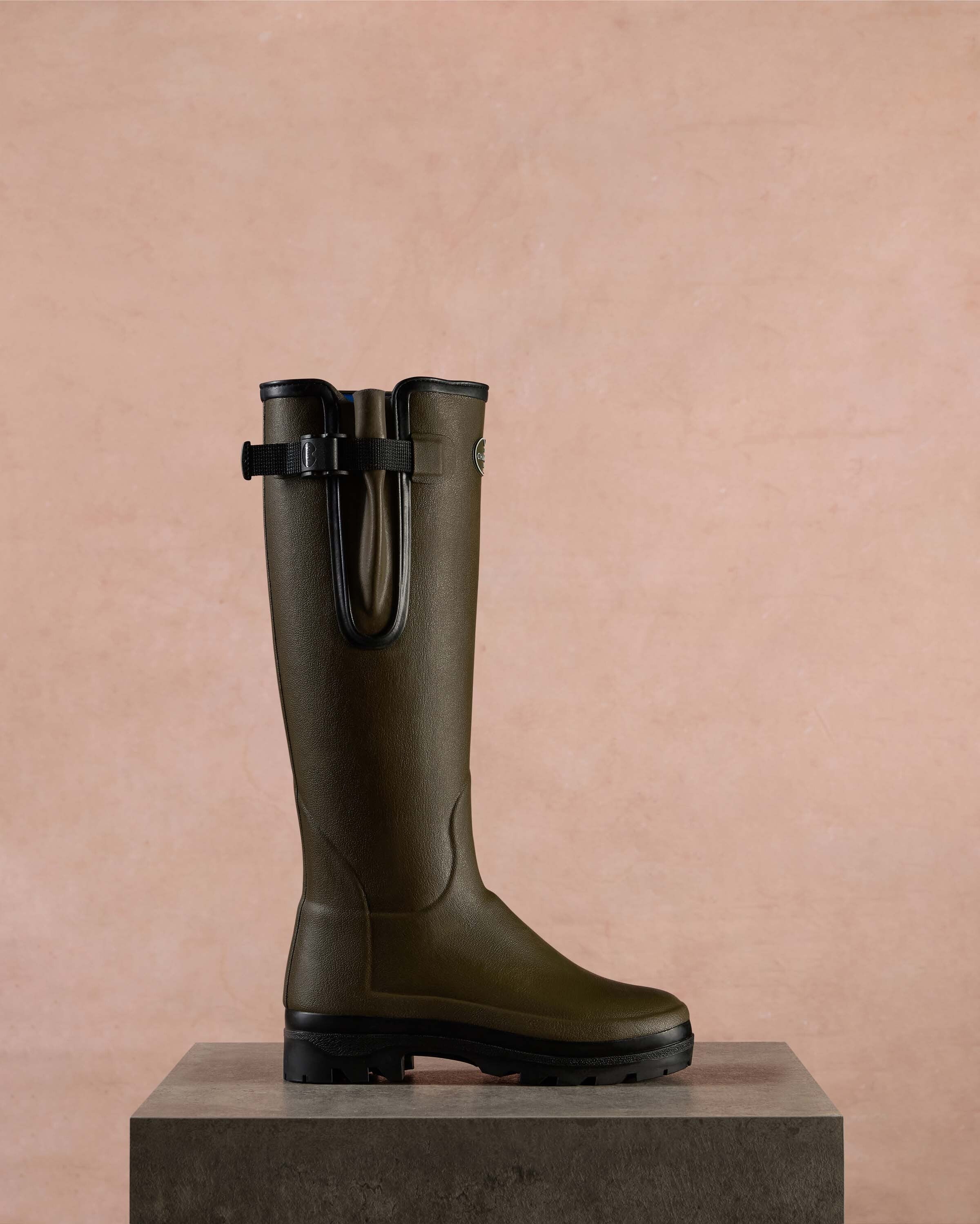 Single Women's Vierzonord Neoprene Lined Wellington Boot in Chameau Green displayed upright on a stone pedestal against a textured blush pink wall. The image focuses on the side profile, showcasing the clean lines, matte finish, and secure strap-and-gusset design.