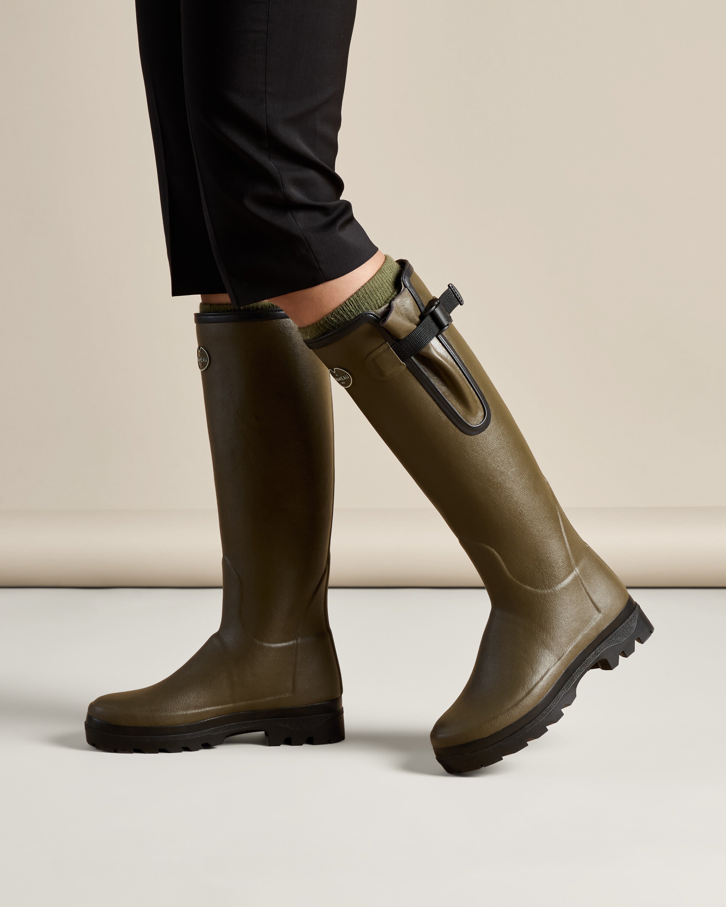 Side view of the Women's Vierzonord Neoprene Lined Wellington Boot in Chameau Green worn by a model mid-stride. The image captures the contrast between the matte green rubber boot body, black sole, and the textured stretch gusset, portraying flexibility and function in motion.